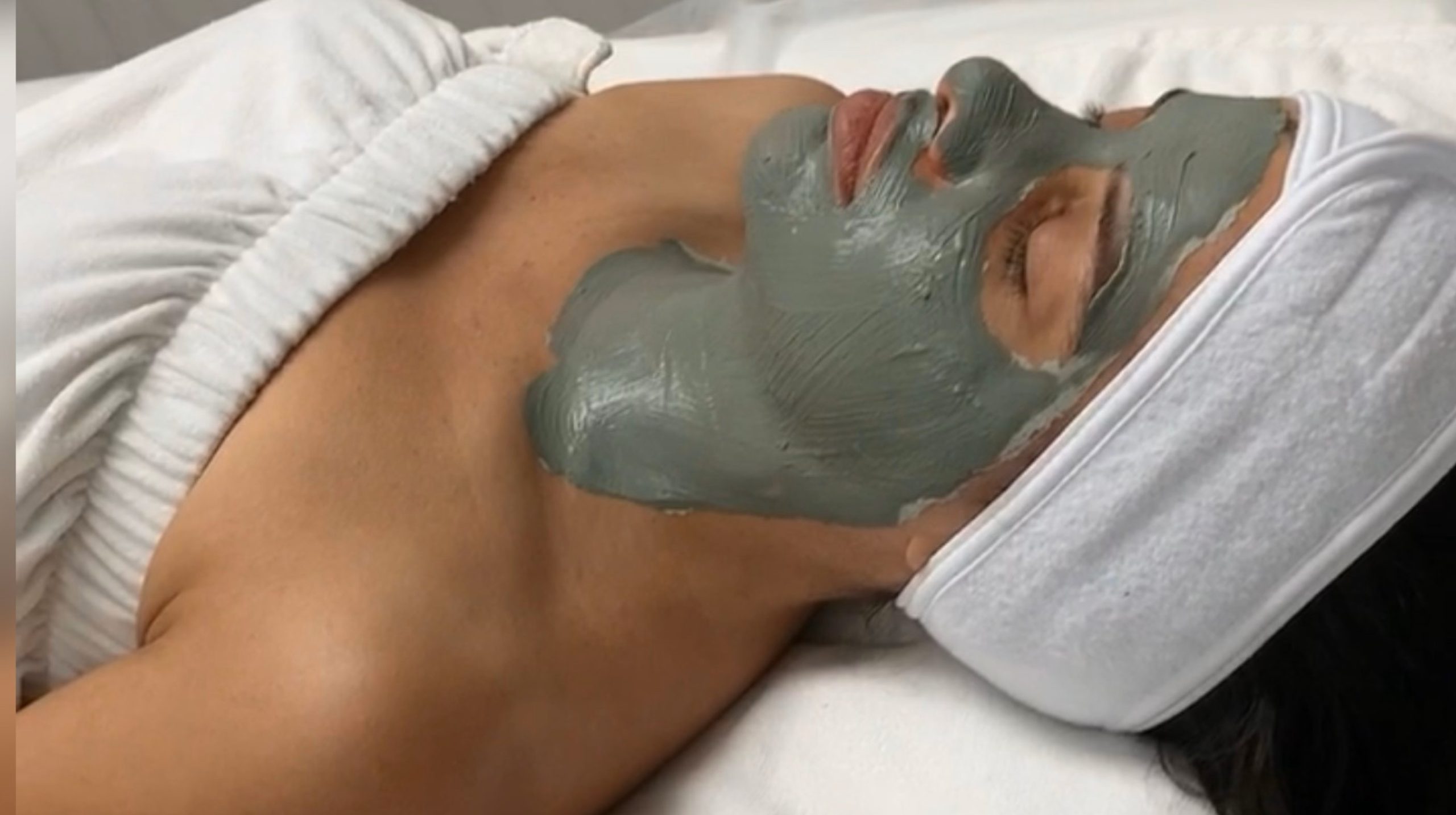 A person lying down receiving a facial treatment at Via Skincare with a thick, grey clay mask applied to their face, excluding the eyes and mouth areas, in a serene spa environment.